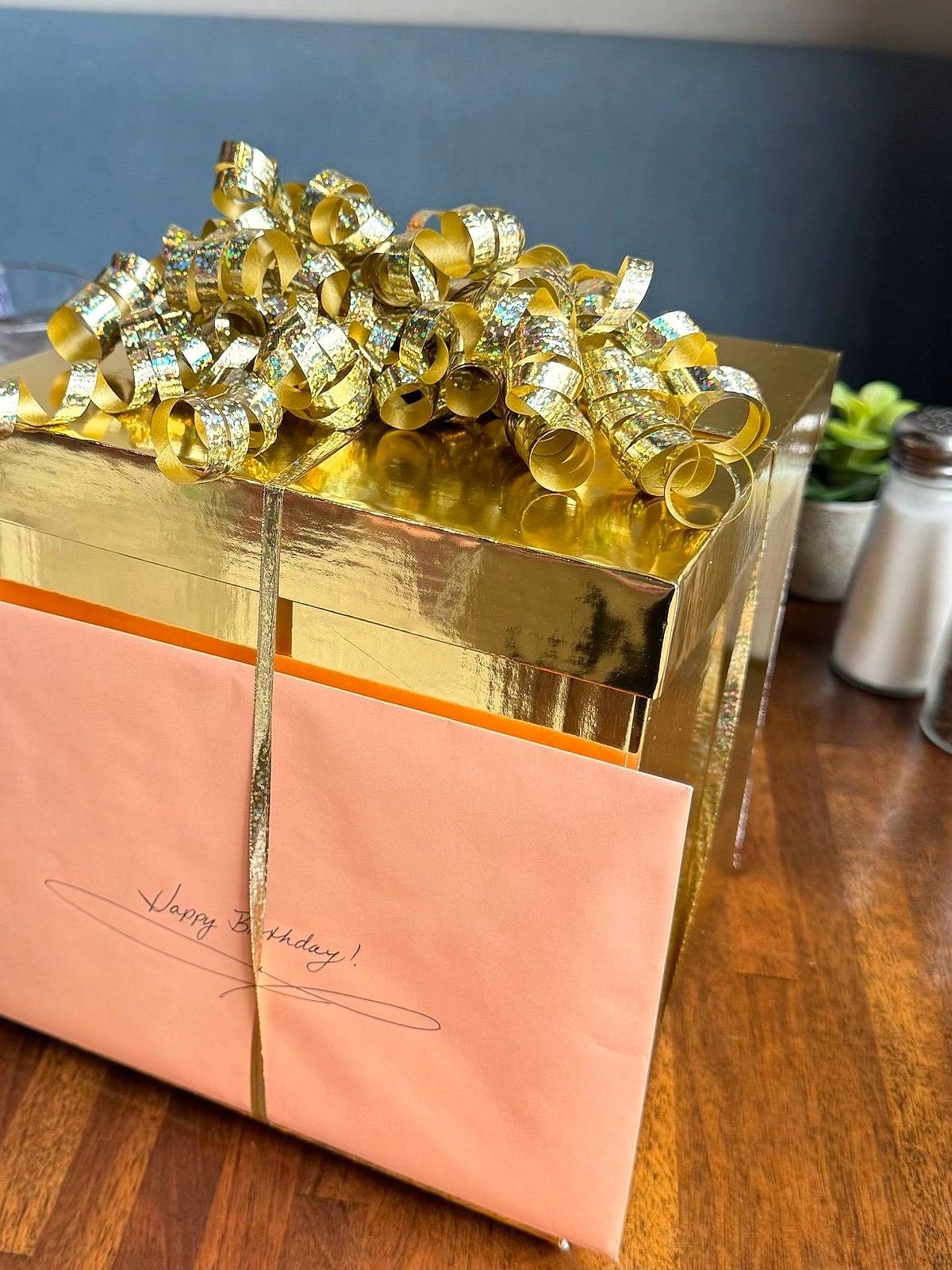 Birthday gift box in shiny gold and peach paper, topped with a cascade of gold curly ribbon